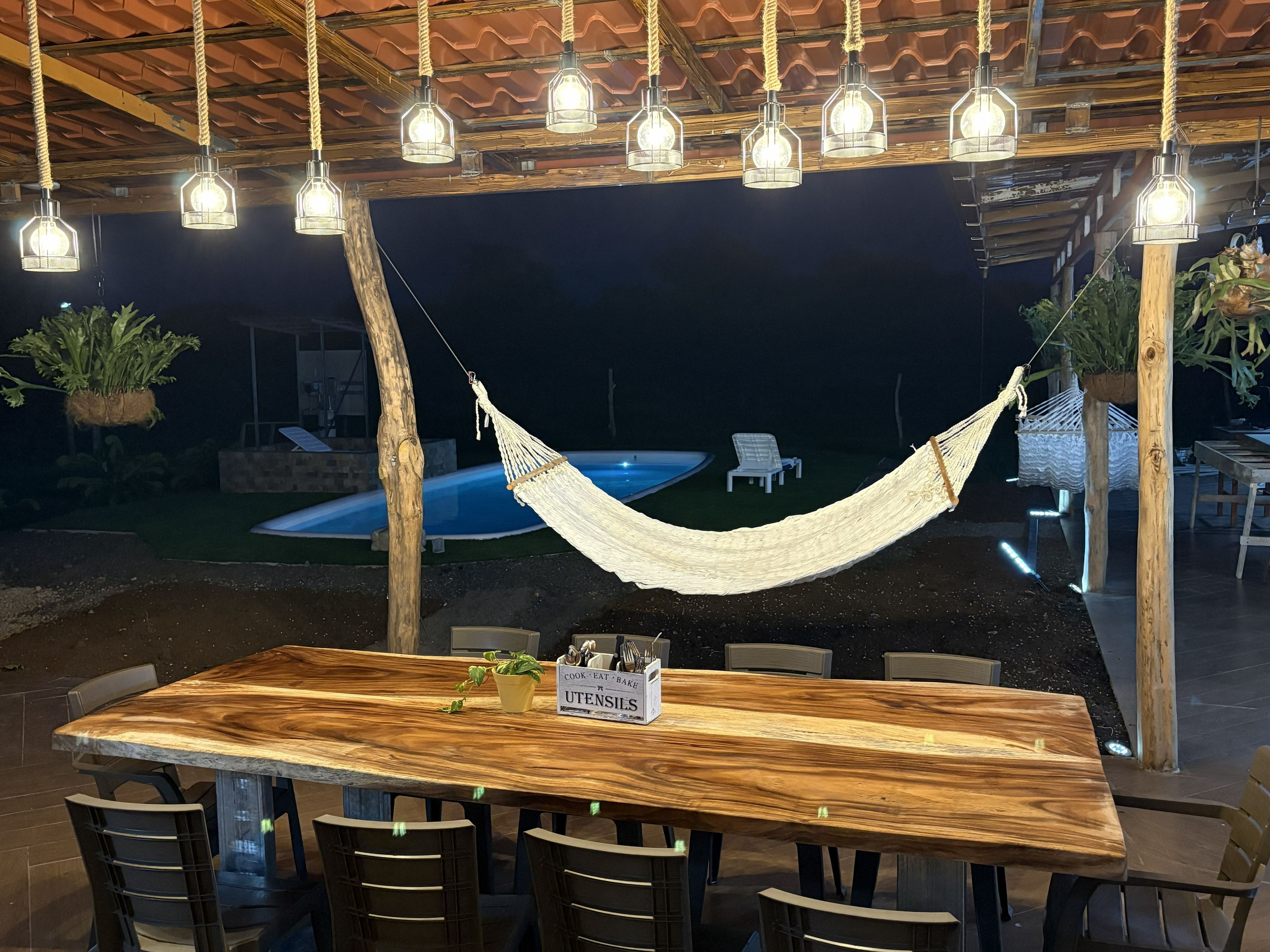 Un espacio de comedor al aire libre con una mesa y sillas de madera, rodeado de lámparas colgantes.