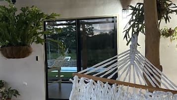 Uma rede com capa de macramé branco, um vaso com planta e vista para uma piscina através de uma porta de vidro.