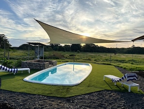 A swimming pool with a sunshade, lounge chairs, and a grassy area.