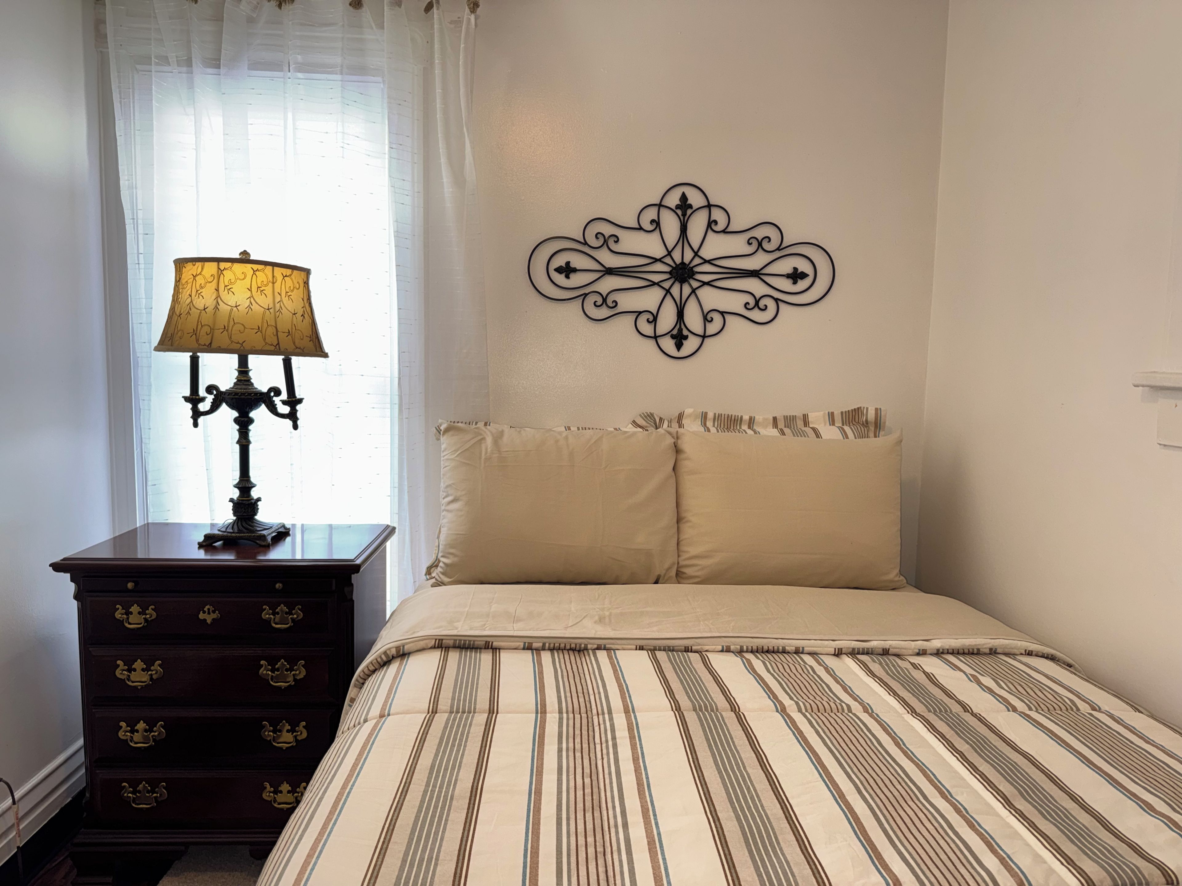 A bedroom with a bed, a nightstand with a lamp, a dresser with drawers, and a decorative wall piece.