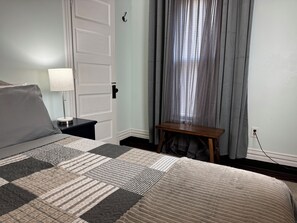 A neatly made bed with a patterned bedspread, a bedside lamp, a small wooden table, and a window with sheer curtains.