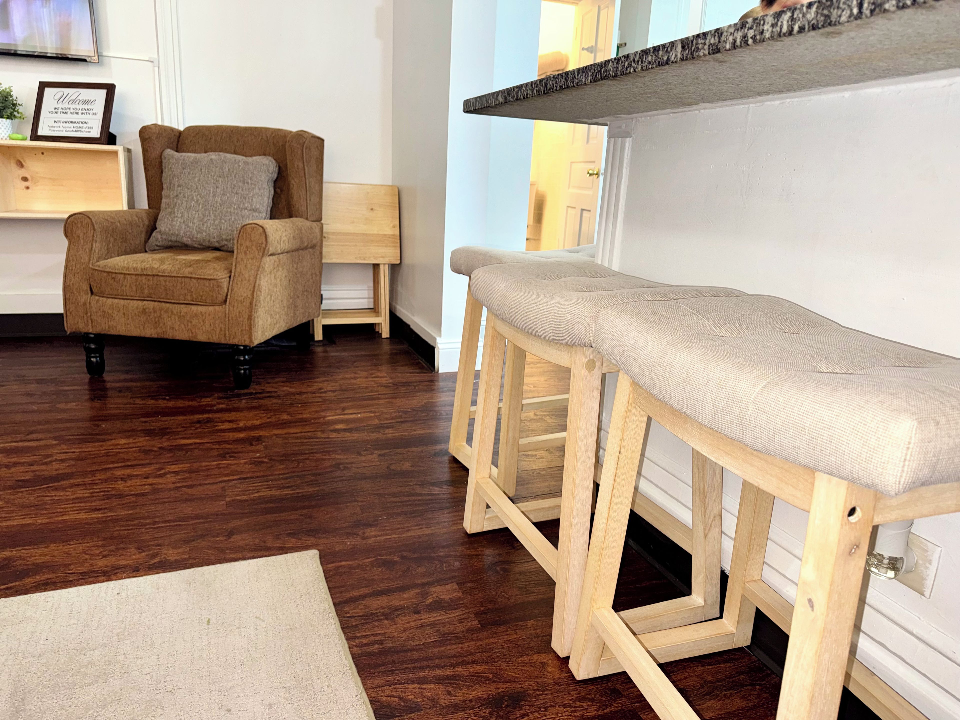 A wooden bar stool with a cushioned seat, a brown upholstered chair with a pillow, and a wooden side table with a framed sign.