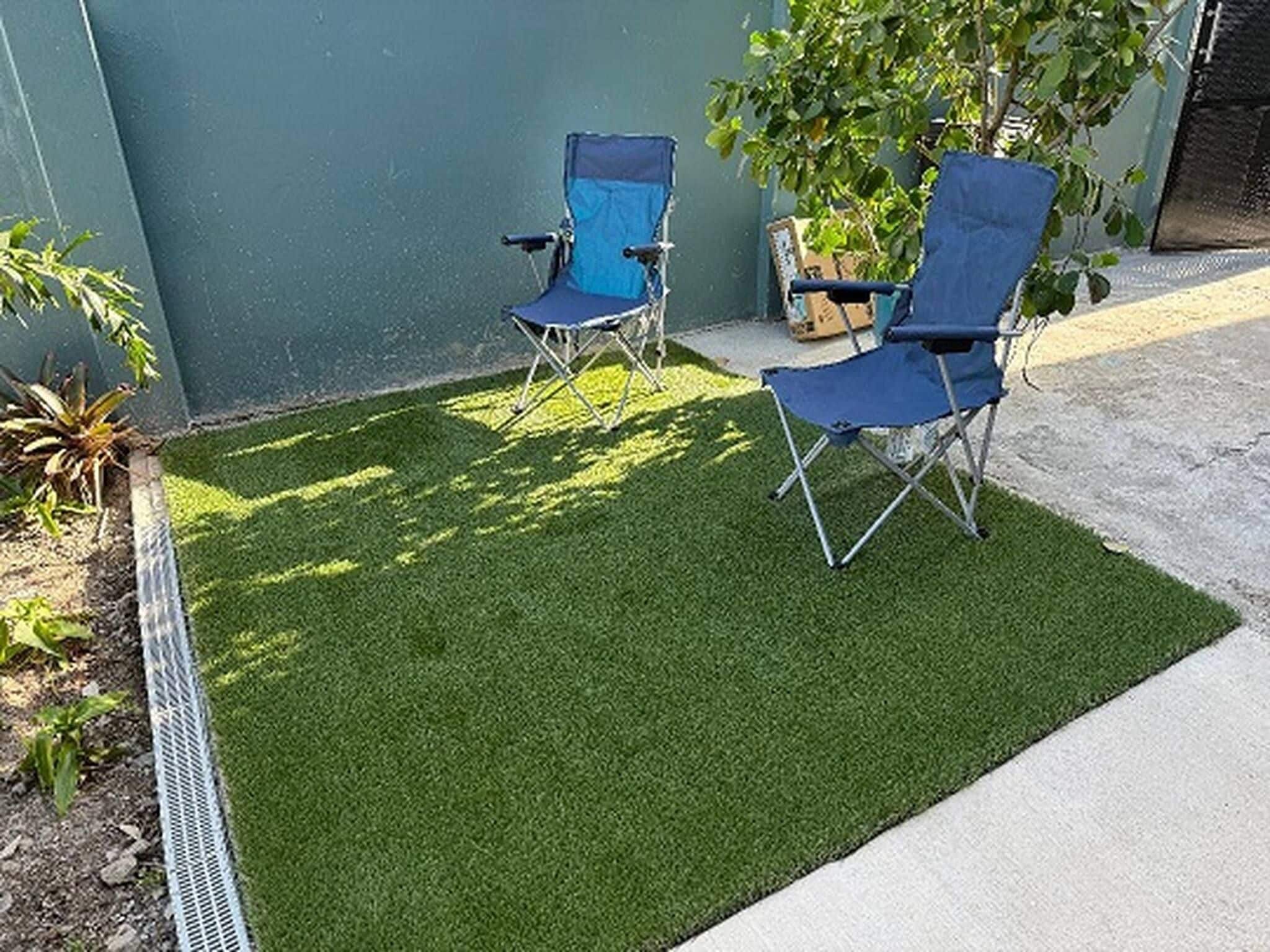 Un petit espace extérieur avec du gazon synthétique, deux chaises pliantes bleues et un pot de fleurs.
