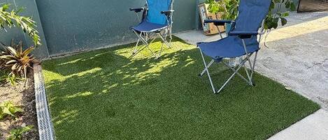 Un petit espace extérieur avec du gazon synthétique, deux chaises pliantes bleues et un pot de fleurs.