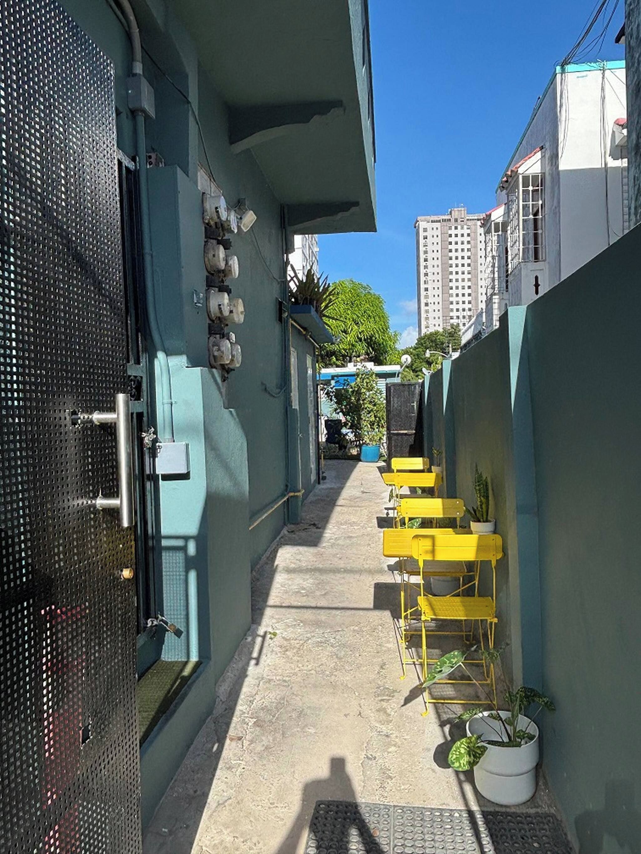 A narrow alleyway with a blue wall, a metal gate, and yellow chairs with potted plants.