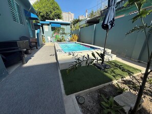 Un patio trasero residencial con piscina, zona de estar en la terraza y un pequeño jardín.
