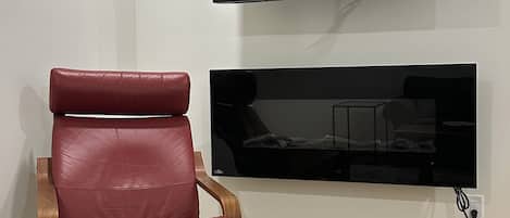 Habitación con televisor de pantalla plana montado en la pared, un sillón de cuero, una mesa de madera y un control remoto sobre la mesa.