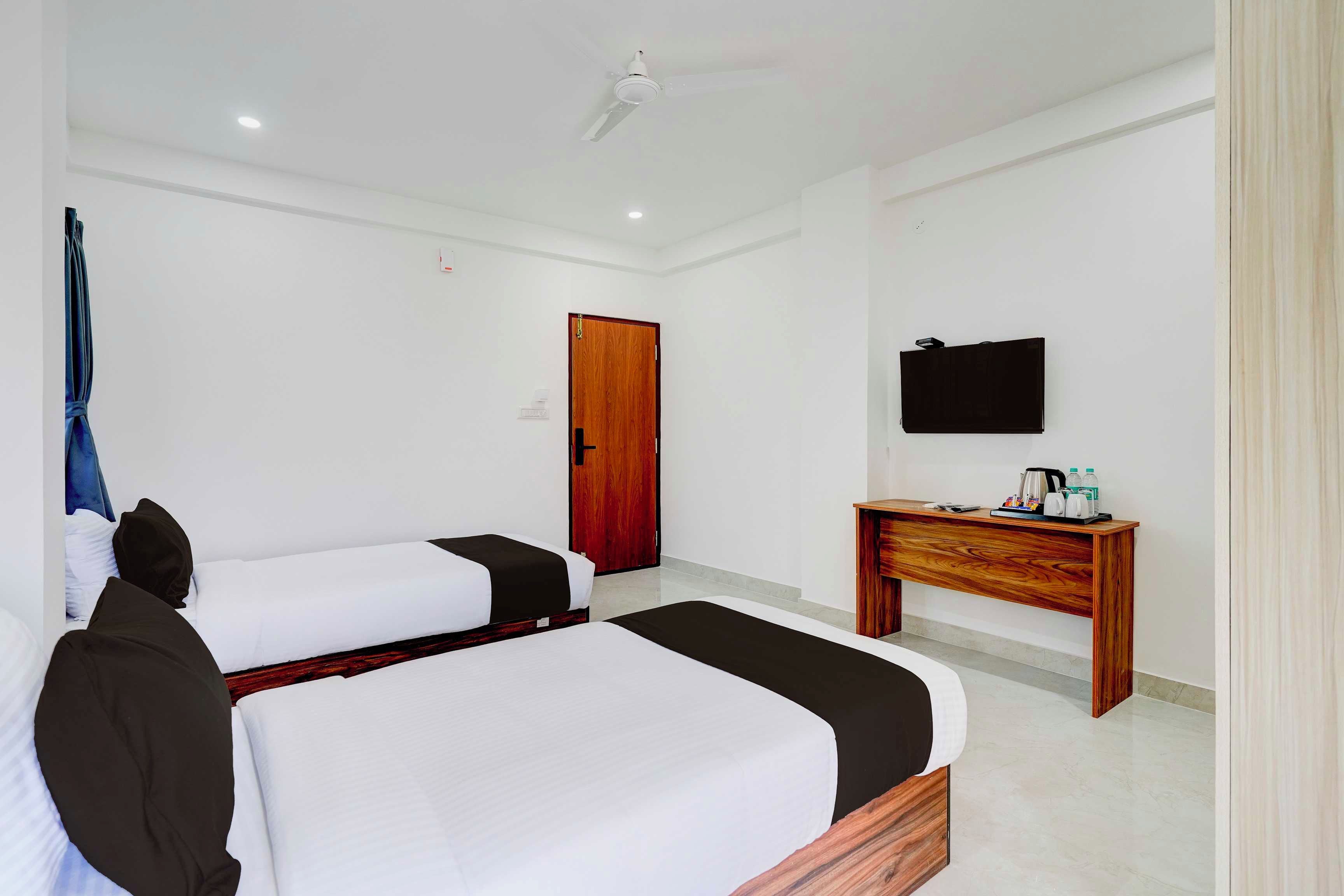 A hotel room with two beds, a wooden door, a TV on the wall, and a small wooden table with a kettle and bottles.