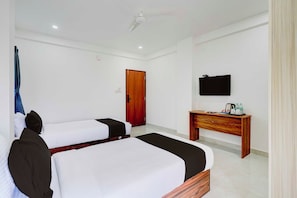 A hotel room with two beds, a wooden door, a TV on the wall, and a small wooden table with a kettle and bottles.