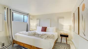 A neatly made bed with a white comforter and a knitted throw, a bedside table with a lamp, and a patterned rug on the floor.