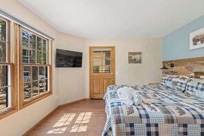 Ein Schlafzimmer mit einem großen Fenster, einem Bett mit gemustertem Überwurf, einer Holztür und einem an der Wand befestigten Fernseher.
