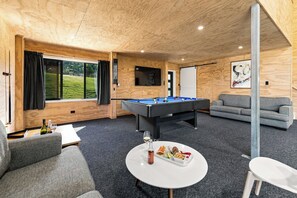A room with a pool table, grey sofa, and a dining table with food and drinks. - The Black Barn - Whakatane Holiday Home (Whakatane)