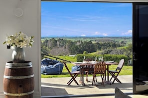 A dining area with a table and chairs, a vase with flowers, and a view of a landscape through a glass door. - The Black Barn - Whakatane Holiday Home (Whakatane)
