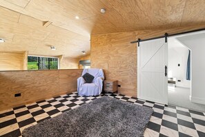 Una habitación con suelo de cuadros, un alfombra de pelo alto gris, una silla blanca con almohada y un techo de madera.