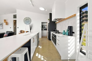 A modern kitchen with white cabinetry, a central island, and a view through a window. - The Black Barn - Whakatane Holiday Home (Whakatane)