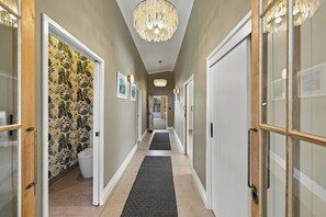 A hallway with a patterned wallpaper, a chandelier, and a black rug leading to a door. - The Black Barn - Whakatane Holiday Home (Whakatane)