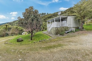 A single-story house with a porch, surrounded by a grassy area and trees. - Treetops & Tide Cottage - Moetapu Holiday Home (Picton)