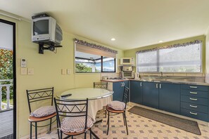 A compact kitchen with a dining area, a television mounted on the wall, and a window with a view of greenery. - Treetops & Tide Cottage - Moetapu Holiday Home (Picton)