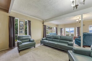A spacious living room with a large sofa, two armchairs, and a view of the outdoors through large windows. - The Bay House - Moetapu Holiday Home (Picton)