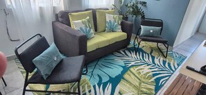 Ein Wohnzimmer mit einem grauen Sofa, einem gelben Kissen, einem blau-grün gemusterten Teppich und einer Topfpflanze.