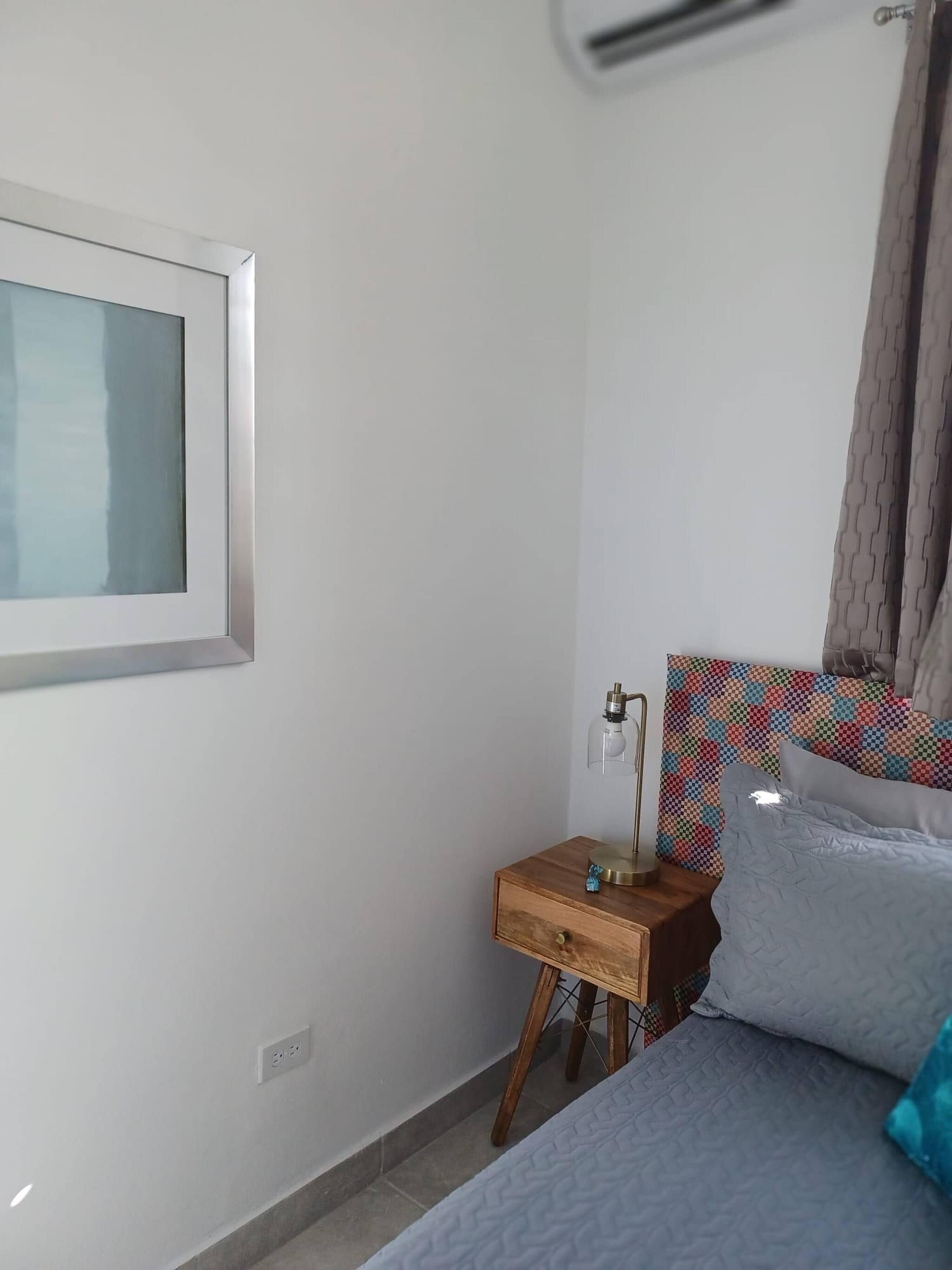 A bedroom with a bed, a bedside table with a lamp, a window, and a colorful headboard.