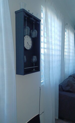 Una habitació amb un rellotge penjat a la paret, una finestra amb cortines transparents i un llit.