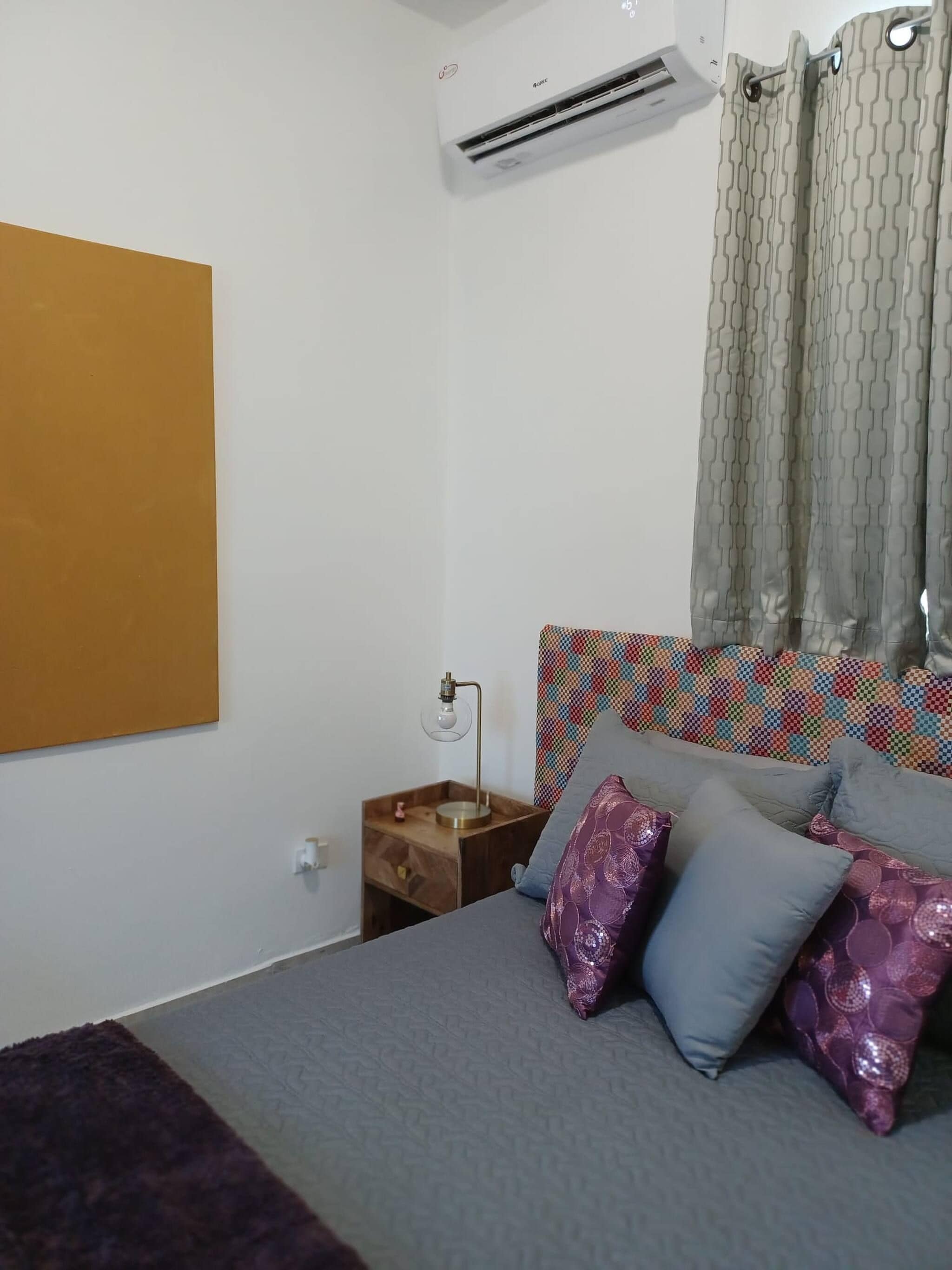 A bedroom with a bed, a nightstand, an air conditioning unit, and a patterned curtain.