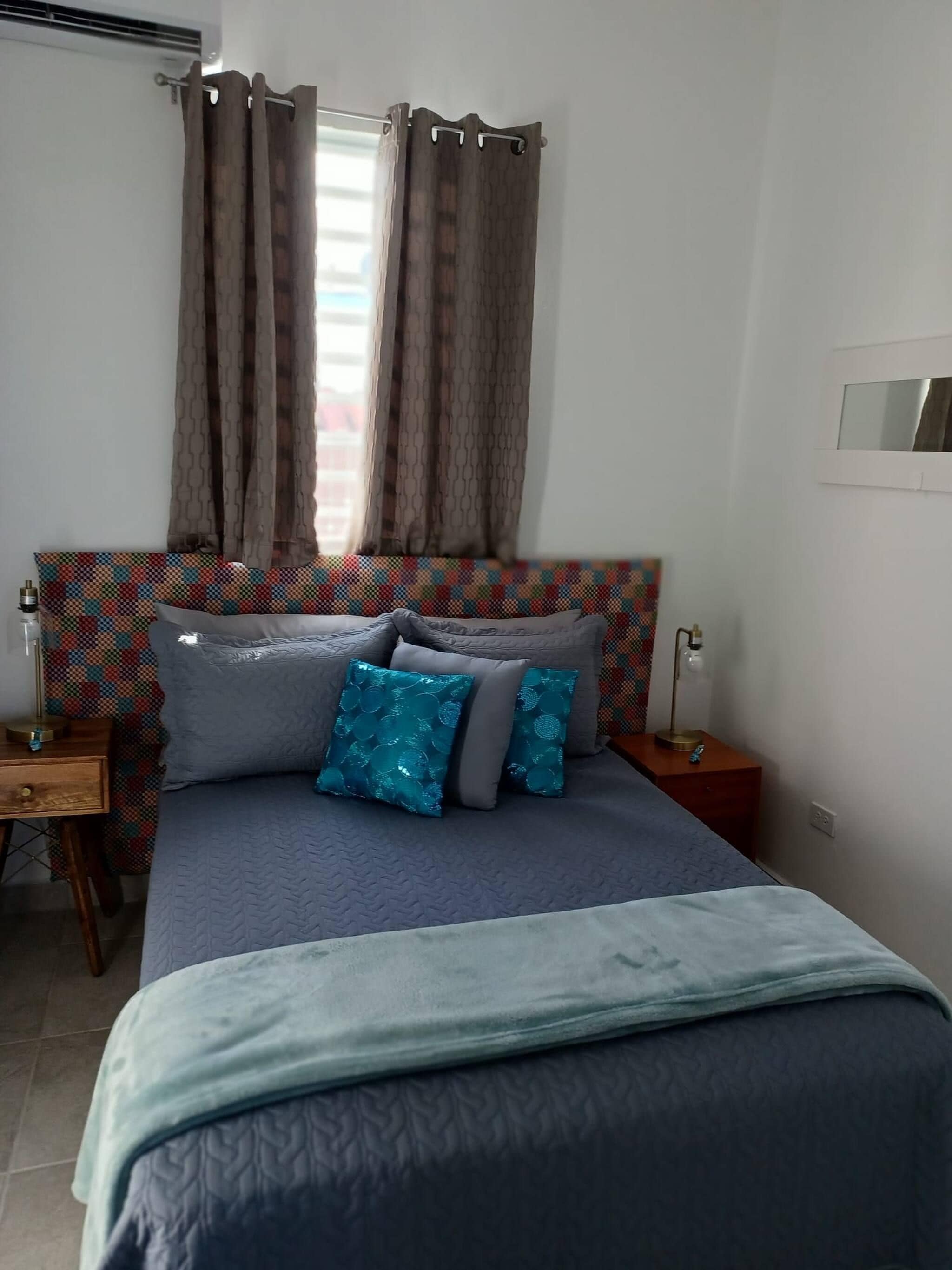Quarto com cama, mesa de cabeceira, luminária e janela com cortinas.