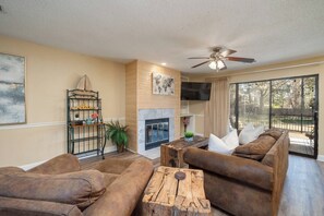 A living room with a fireplace, a ceiling fan, and a large window with a view of the outdoors. - Emerald Isle Condo - 2bed/2bath Lots of Amenities! (Hot Springs)
