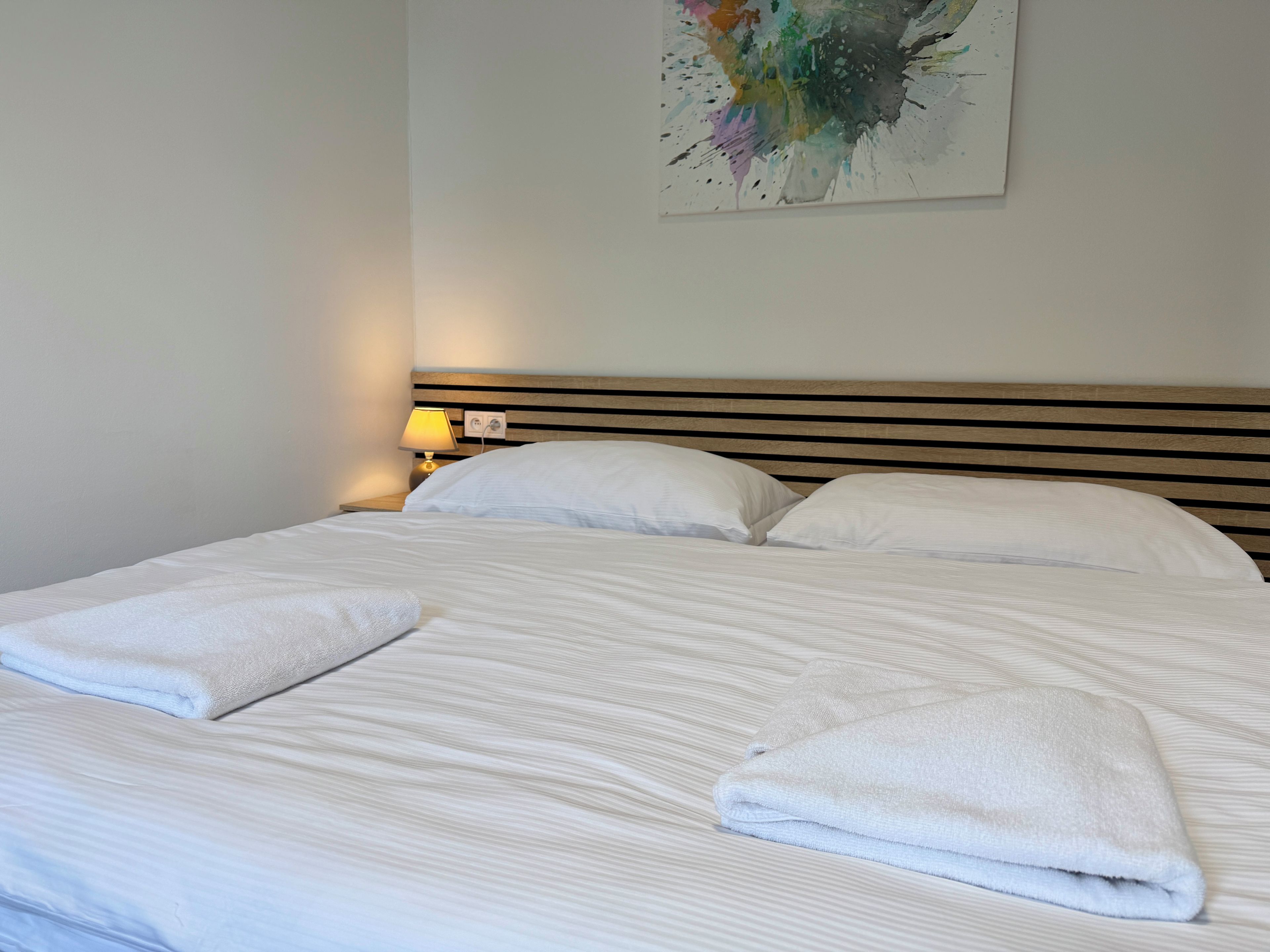 A neatly made bed with white linens and two folded towels, a bedside lamp, and a colorful abstract painting on the wall.