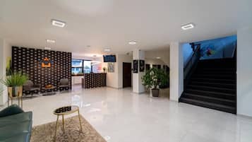 Un vestíbulo de hotel con mostrador de recepción, zona de estar, escalera y plantas en macetas.
