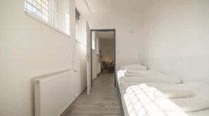 Un pasillo estrecho con un radiador a la izquierda, una cama con ropa blanca a la derecha y una puerta que da a otra habitación.