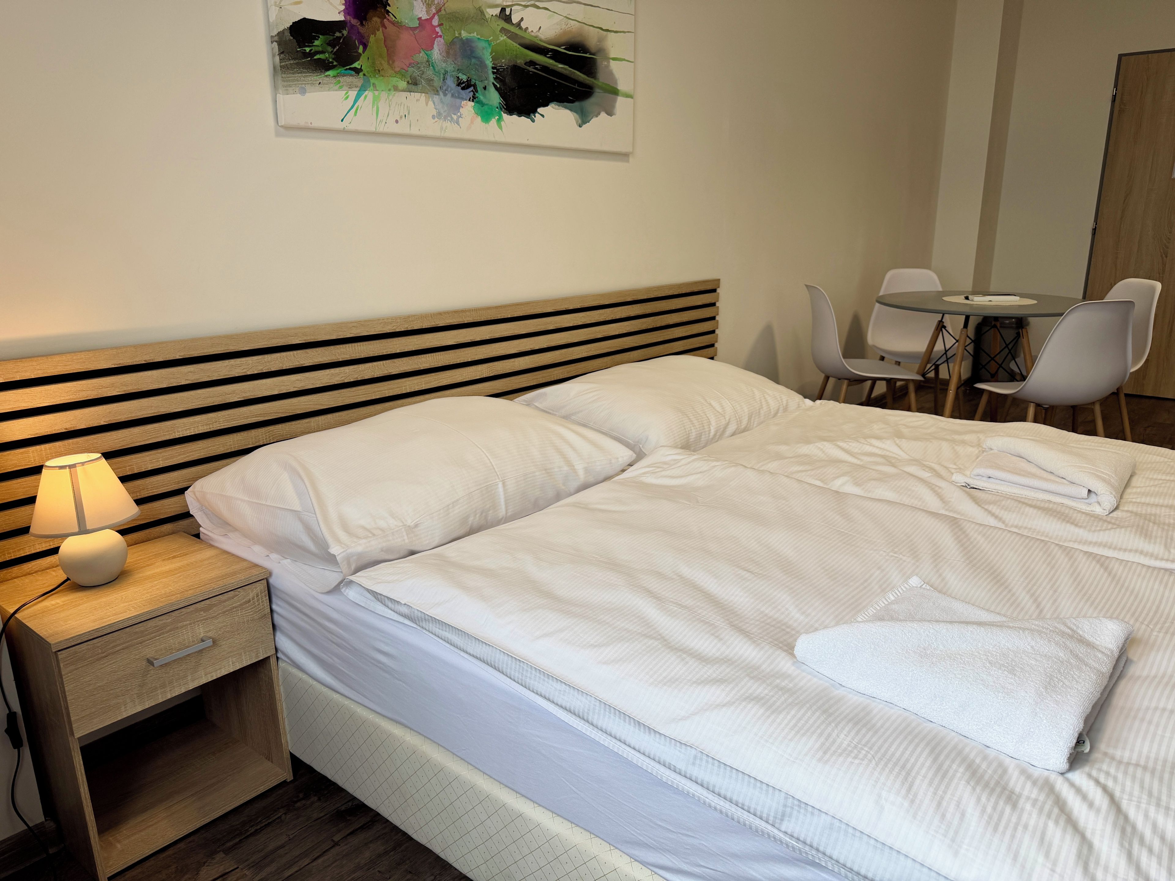 A neatly made bed with pillows, a bedside table with a lamp, and a chair at a small table.