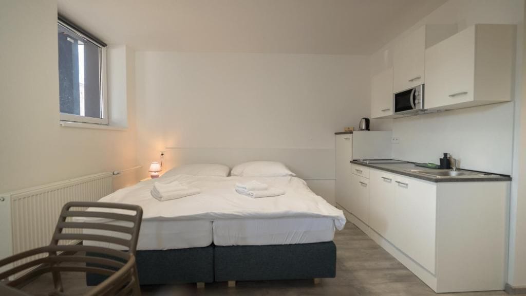 Una habitación pequeña y funcional con cama, una silla, kitchenette y ventana.
