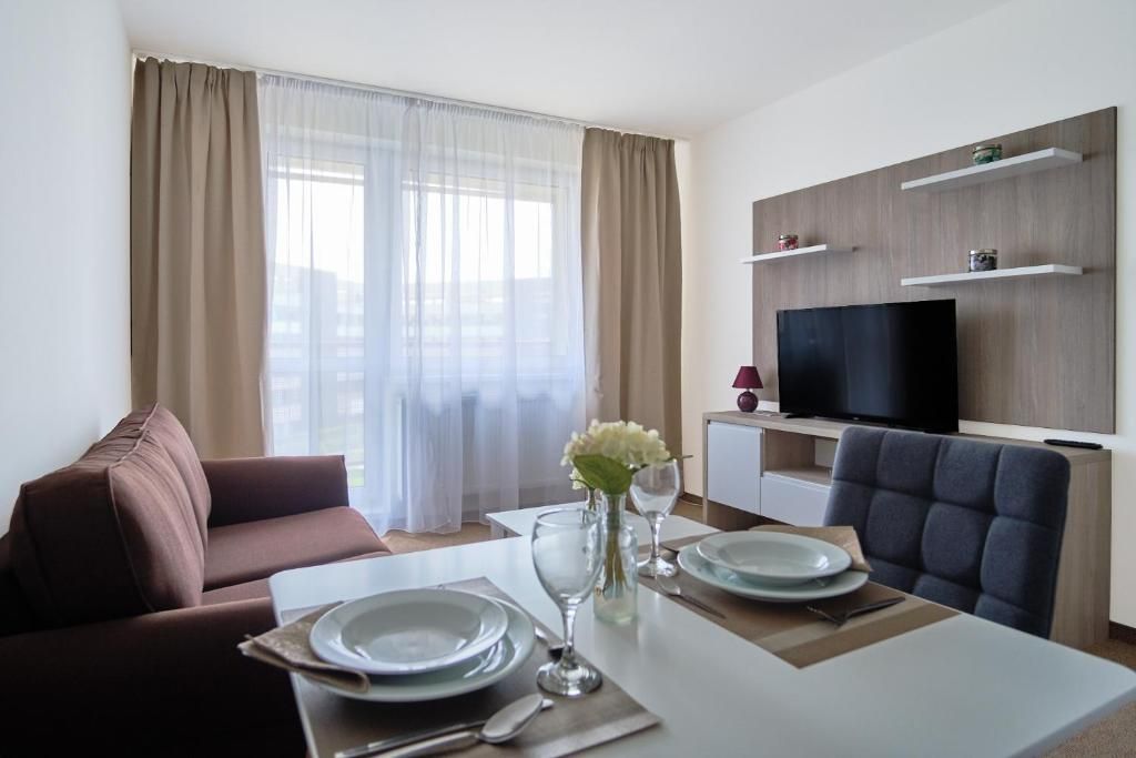 A modern living room with a sofa, dining table set for two, a flat-screen TV, and a window with sheer curtains.