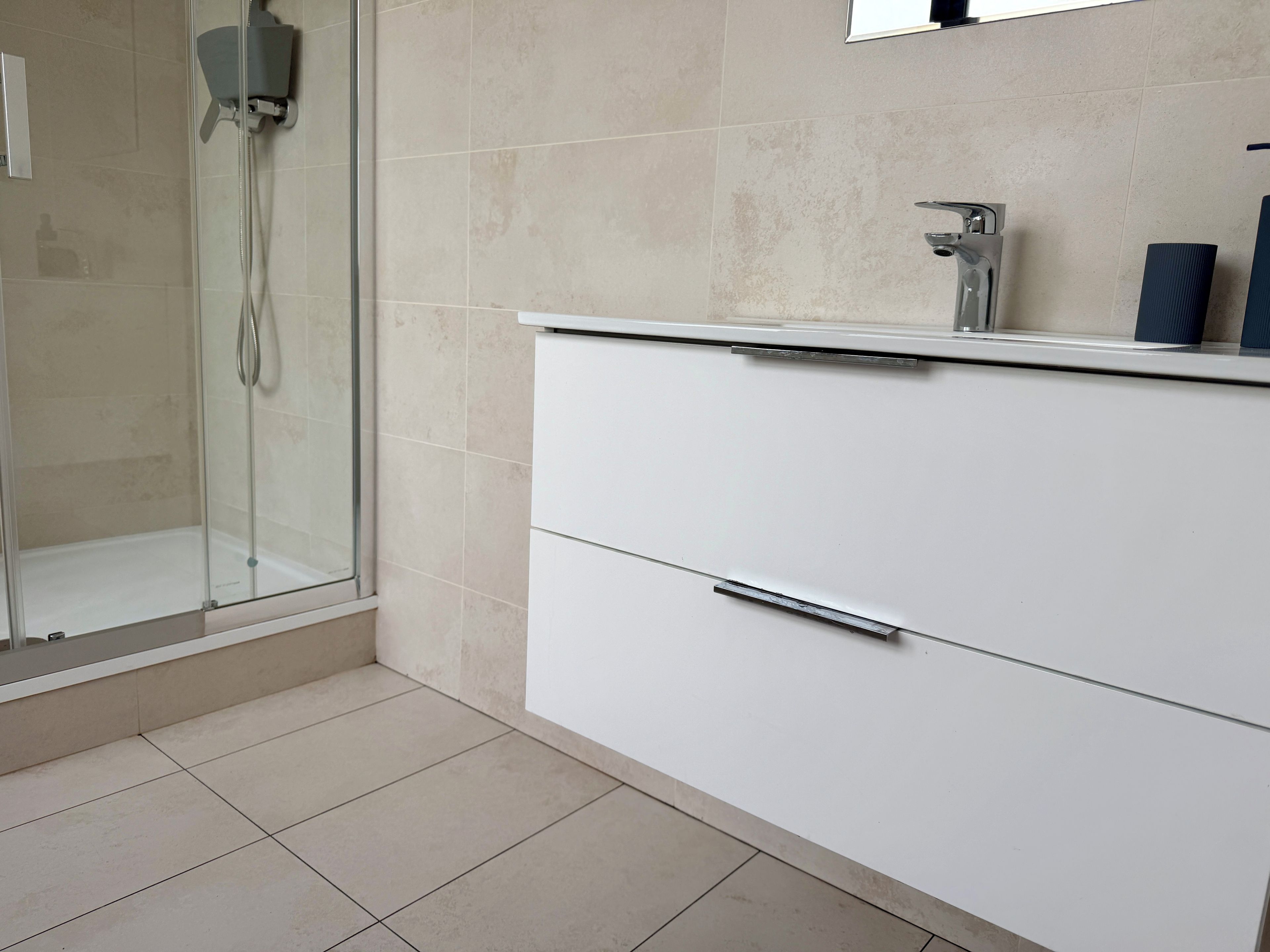 A bathroom with a glass shower enclosure, a white vanity with a mirror, and a tiled floor.