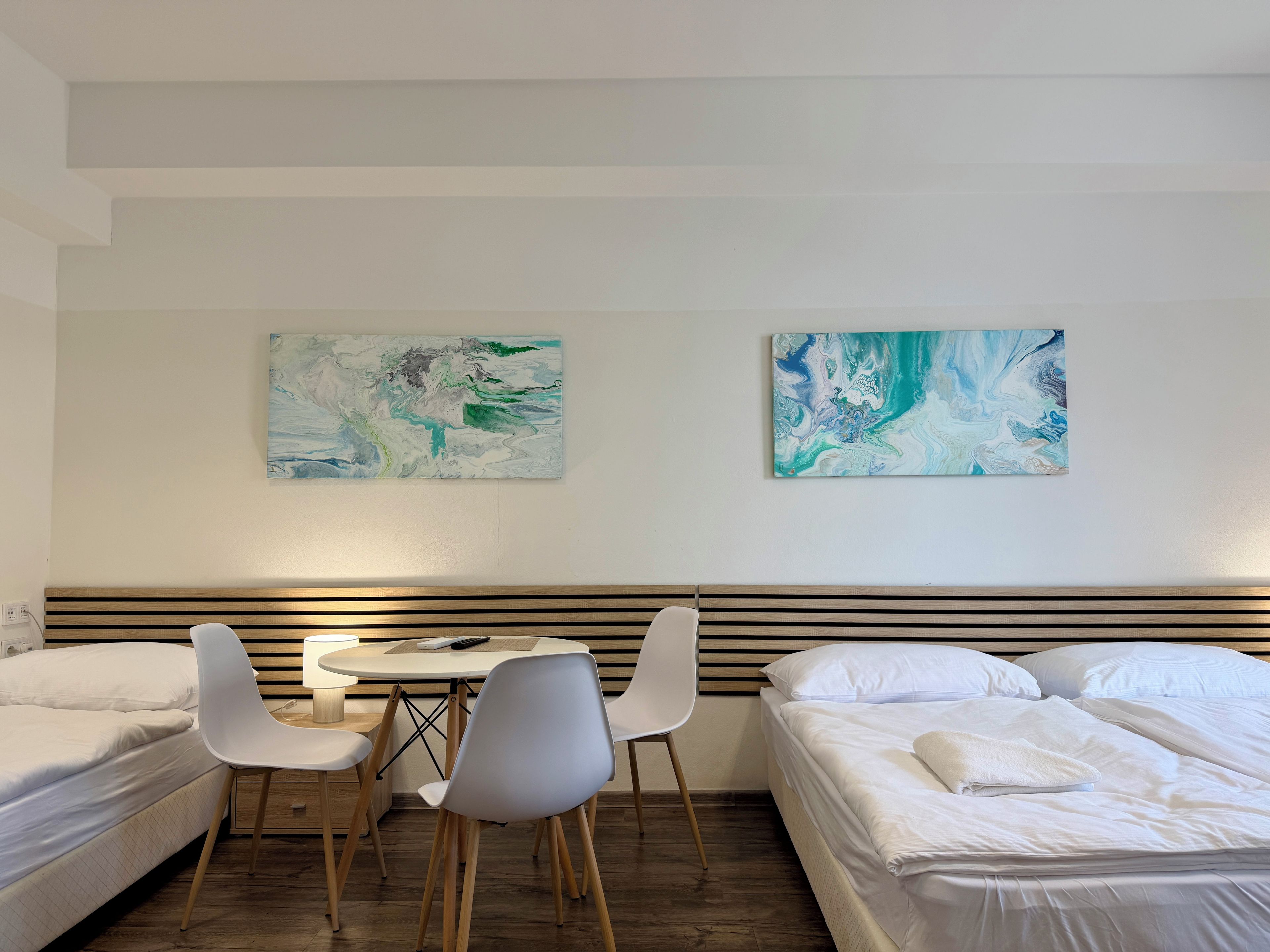 A hotel room with two beds, a dining table, and two chairs. There are abstract paintings on the wall.