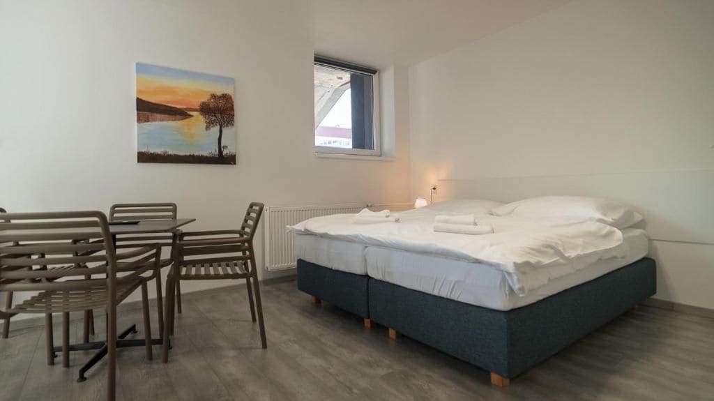 A bedroom with a bed, a desk, chairs, a window, and a painting on the wall.