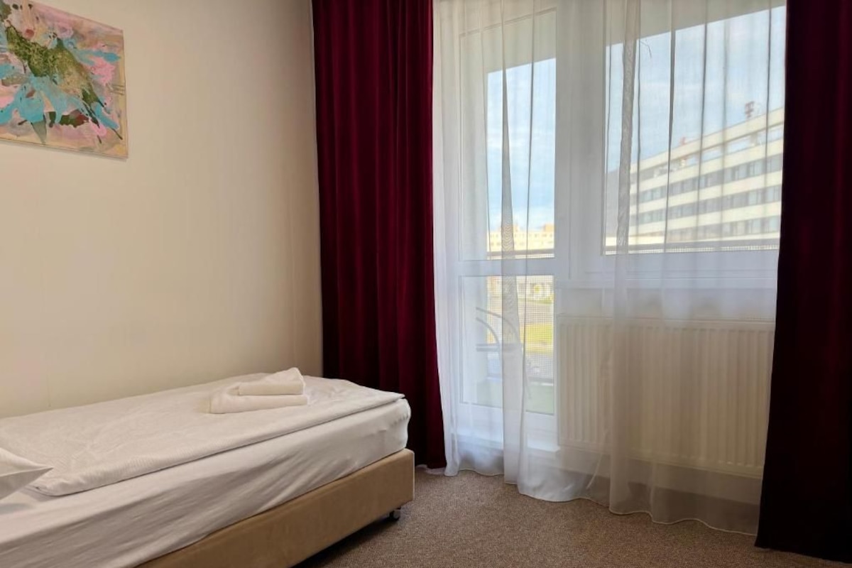 Ein Einzelbett mit weißer Matratze und braunem Kopfteil in einem Zimmer, aus dem ein Blick auf ein Gebäude durch transparente Vorhänge möglich ist.