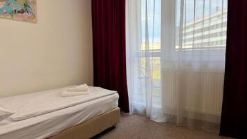 A single bed with a white mattress and a brown headboard in a room with a view of a building through sheer curtains.