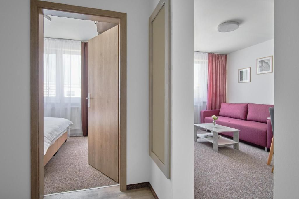 Una habitación de hotel con una cama, una puerta de madera, un sofá rosa, una mesa de centro blanca y un cuadro en la pared.