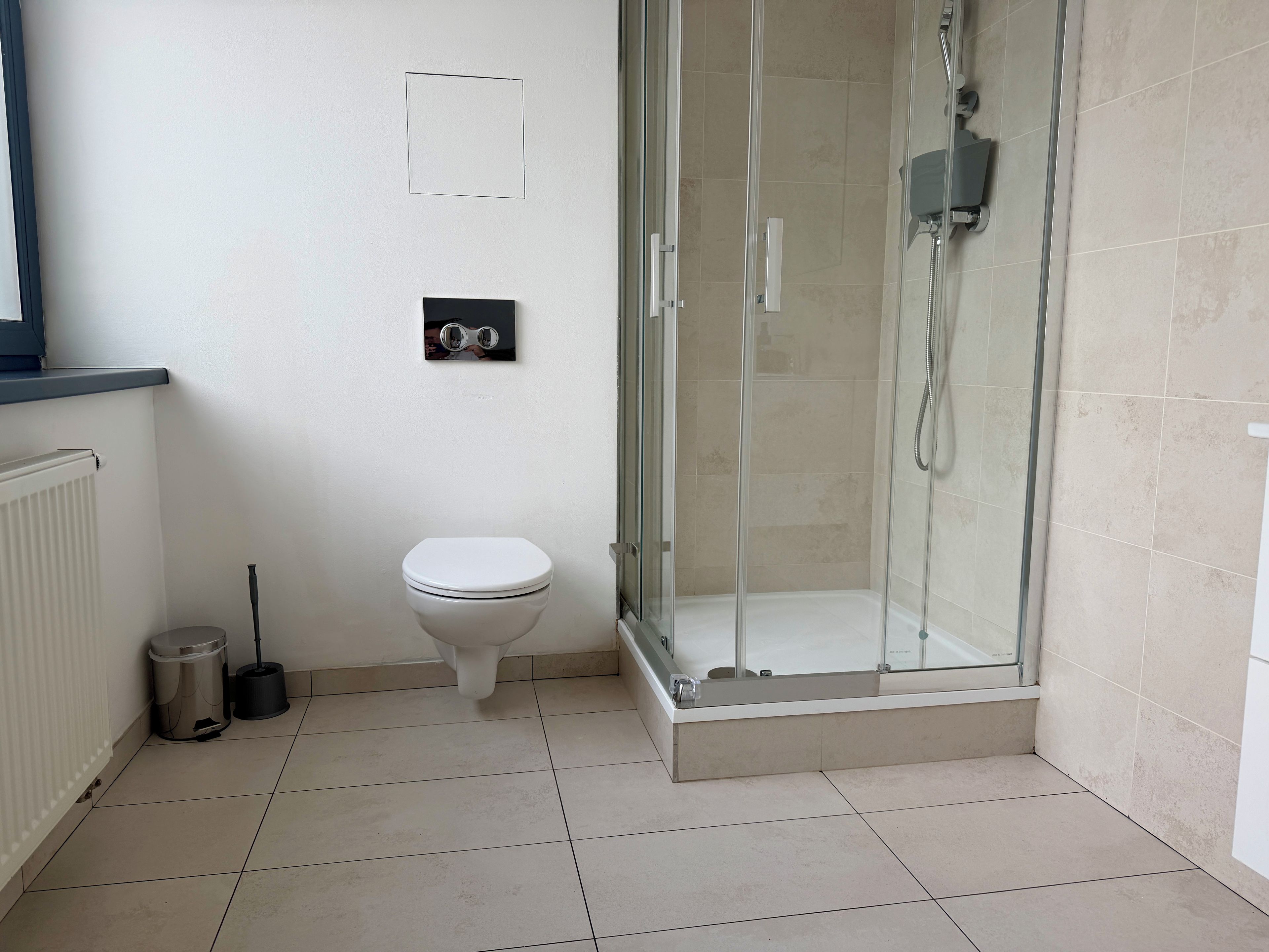 A bathroom with a toilet, a shower enclosure, a radiator, and a trash bin.