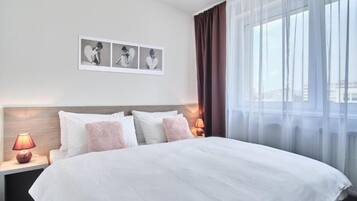 Una habitación de hotel con una cama grande, una mesita de noche con lámpara y una ventana con cortinas translúcidas.
