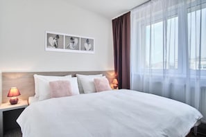 Una habitación de hotel con una cama grande, una mesita de noche con lámpara y una ventana con cortinas translúcidas.
