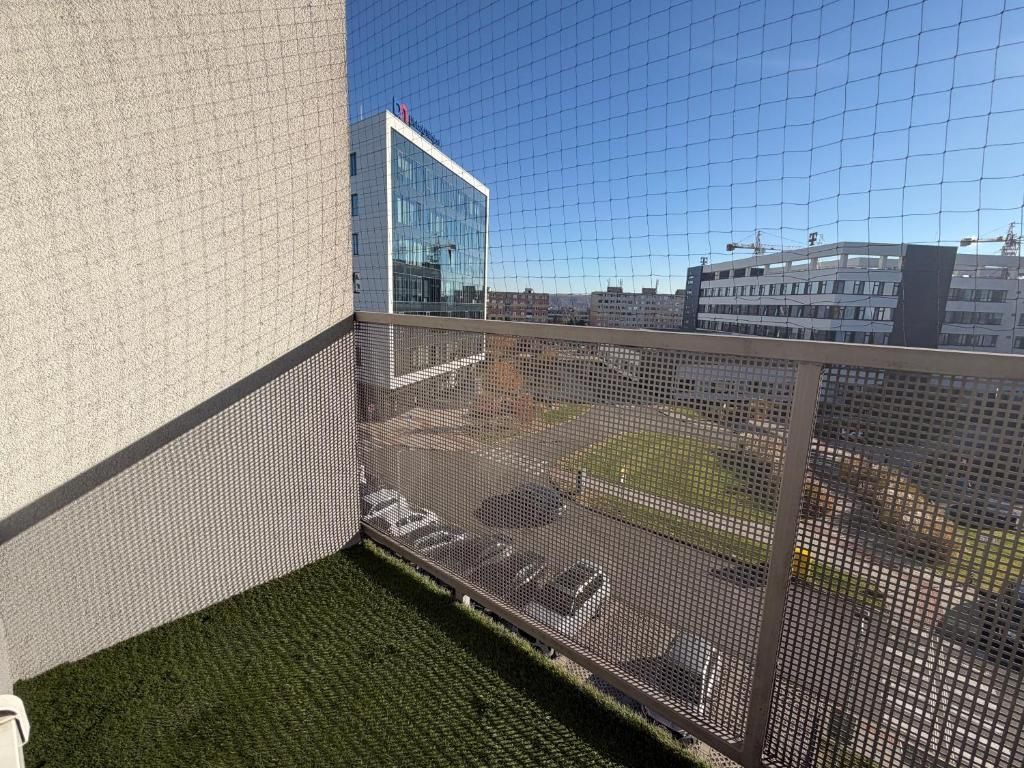 Uma varanda com guarda-corpo de tela, um edifício com fachada de vidro e um estacionamento ao fundo.