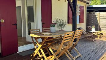 A wooden deck with a table and chairs, a red door, and a white wall with shutters.