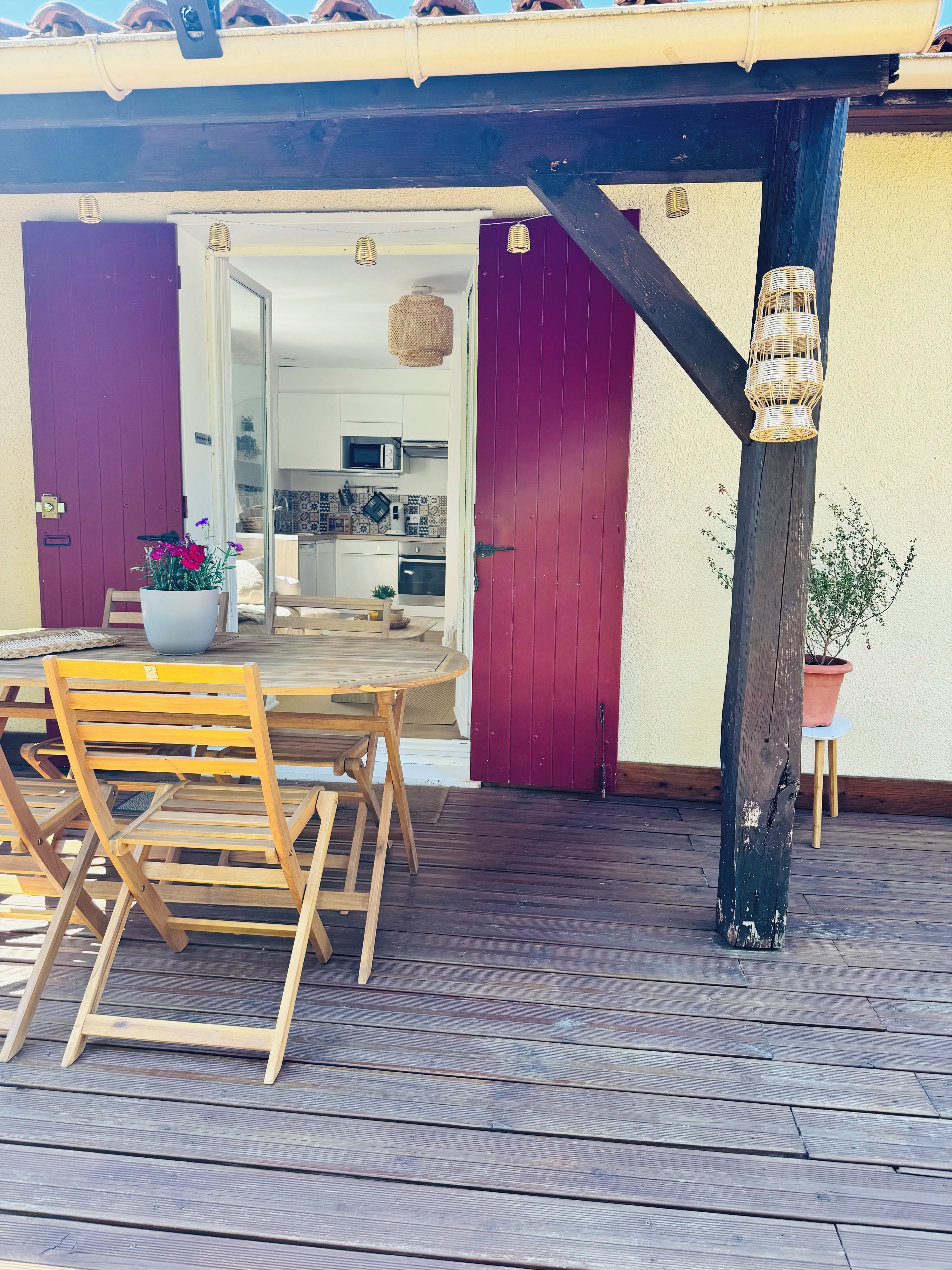Eine Terrasse aus Holz mit Tisch und Stühlen, eine violette Tür und ein durch eine offene Tür einsehbarer Küchenbereich.