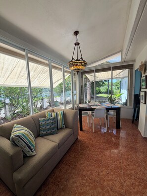 A spacious living area with a sofa, dining table, and chairs. Large windows provide a view of the outdoors. -  Casa BerKana,  Coastal Home, with OCEAN VIEWS, in charming Quebradillas. (San José)