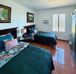 A room with two beds, a nightstand, a lamp, a window with blinds, and a wall-mounted air conditioner. -  Casa BerKana,  Coastal Home, with OCEAN VIEWS, in charming Quebradillas. (San José)