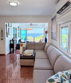 A living room with a large window, a ceiling fan, and a view of the ocean. -  Casa BerKana,  Coastal Home, with OCEAN VIEWS, in charming Quebradillas. (San José)